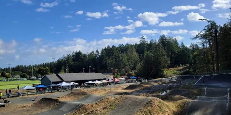 Panoramic view of a BMX track surrounded by forest and a building with tents set up nearby under a blue sky with scattered clouds.