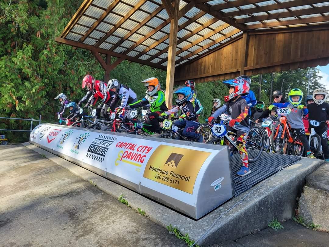 Young BMX riders in racing gear at the starting gate, ready for a race under a covered area with sponsor banners on the front.