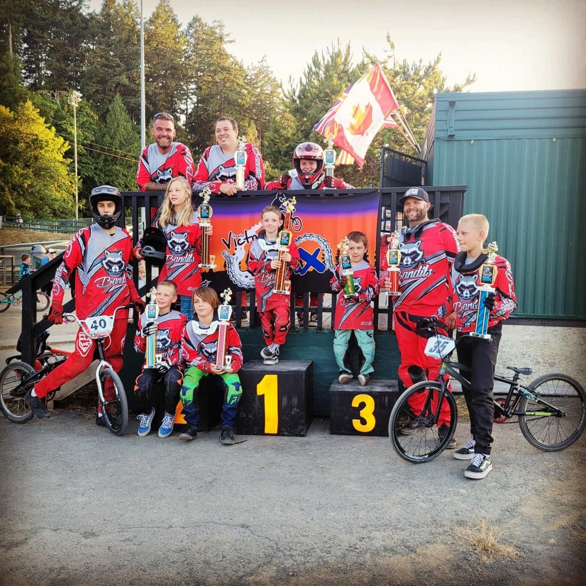 Group of BMX riders in red uniforms posing with their trophies on a podium, with a Canadian flag and a "Victoria BMX" banner in the background.