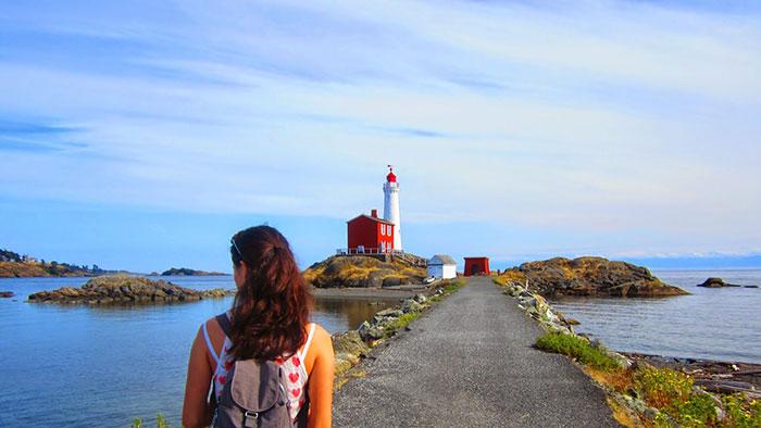 Person with a backpack walking towards the Fisgard Lighthouse, which is situated on a small island connected by a pathway.