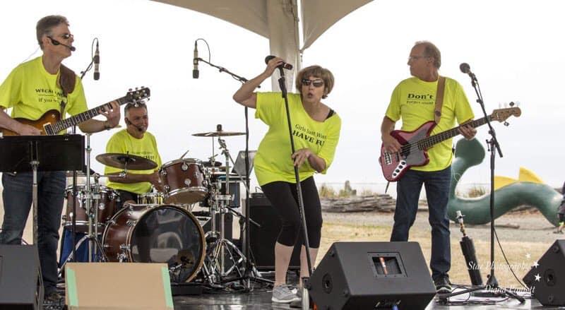 Band performing on outdoor stage, wearing matching bright yellow shirts with humorous slogans.