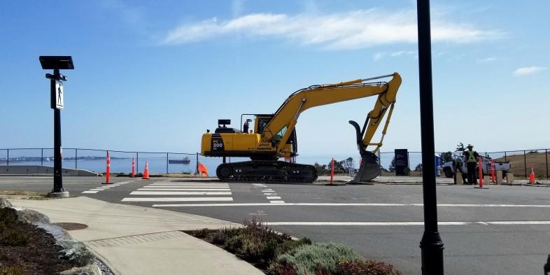 Construction site in Colwood with a large excavator, road work, and a view of the ocean in the background.