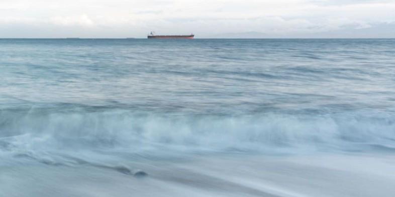 Calm ocean waves with a distant cargo ship on the horizon under a cloudy sky.