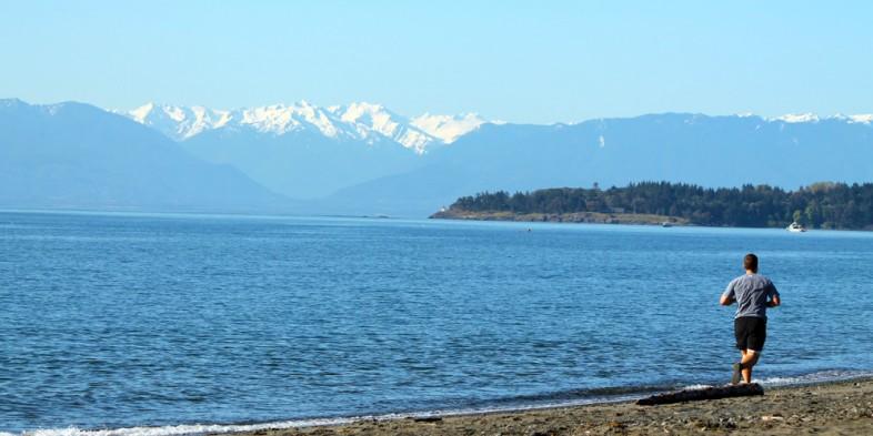 A person jogging on a beach with the sea and snow-capped mountains in the background.