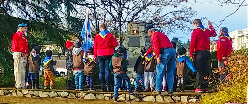 Group of children and adults in scouting uniforms gathered around a monument outdoors.