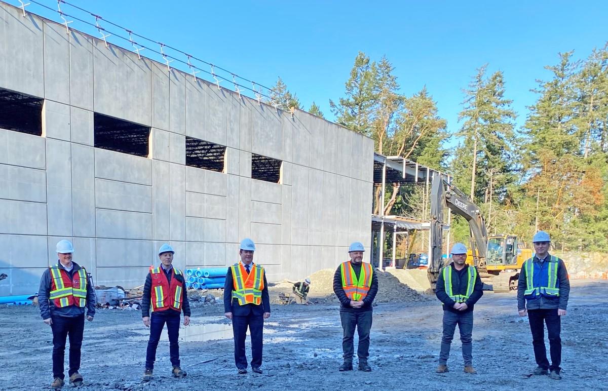 Six construction workers in hard hats and safety vests stand in front of a large building under construction. The site includes an excavator and various building materials.