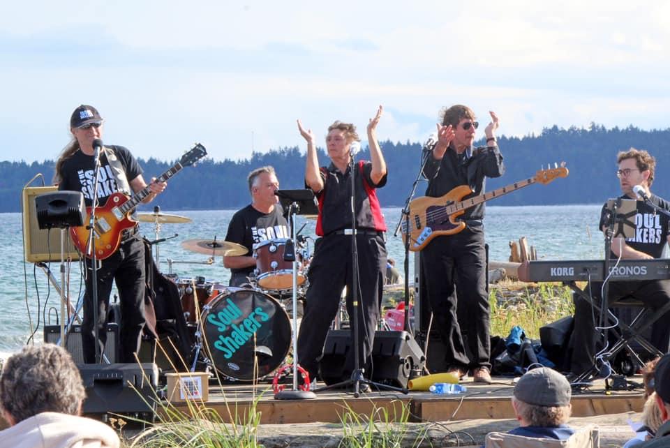 The Soul Shakers band performing outdoors near a beach. The group includes a guitarist, drummer, vocalist, bassist, and keyboardist. They are dressed in black, and an audience watches from the foreground.