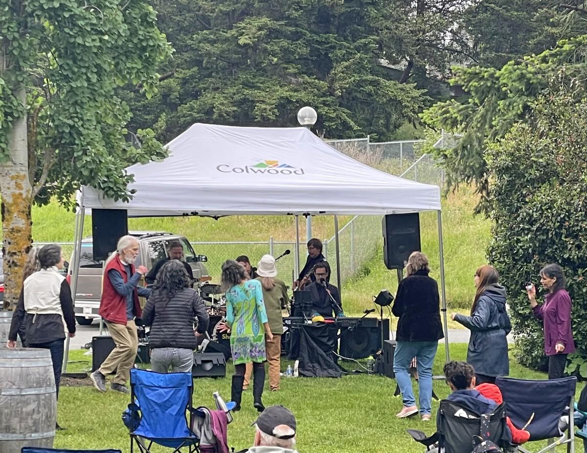 Outdoor event with people gathered under a white tent labeled "Colwood." A band performs under the tent while attendees dance and socialize. The setting is grassy with trees and a fence in the background.