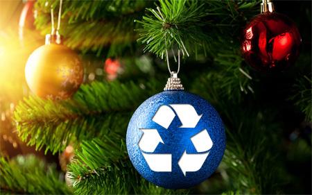 Close-up of a Christmas tree branch adorned with ornaments, including a blue ornament featuring a white recycling symbol, alongside a gold and a red ornament. Sunlight filters through the background.