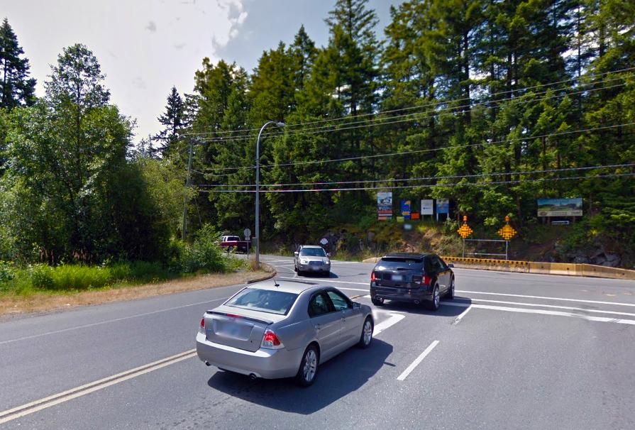 Intersection with several cars stopped and waiting to turn, surrounded by dense trees and greenery. Signs and road markings are visible, with power lines crossing overhead. The scene is brightly lit under a clear sky.