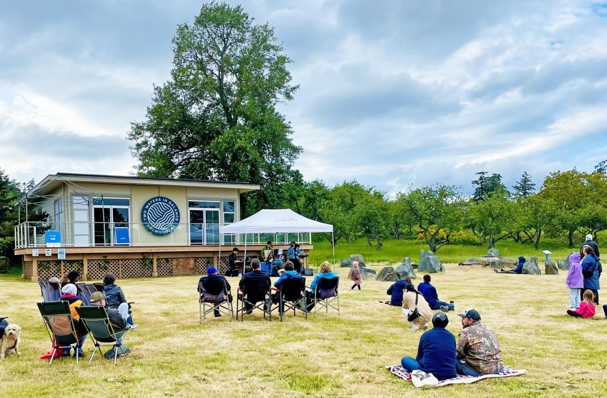 Outdoor event with people seated on the grass facing a small building, with a stage and canopy tent set up in front.